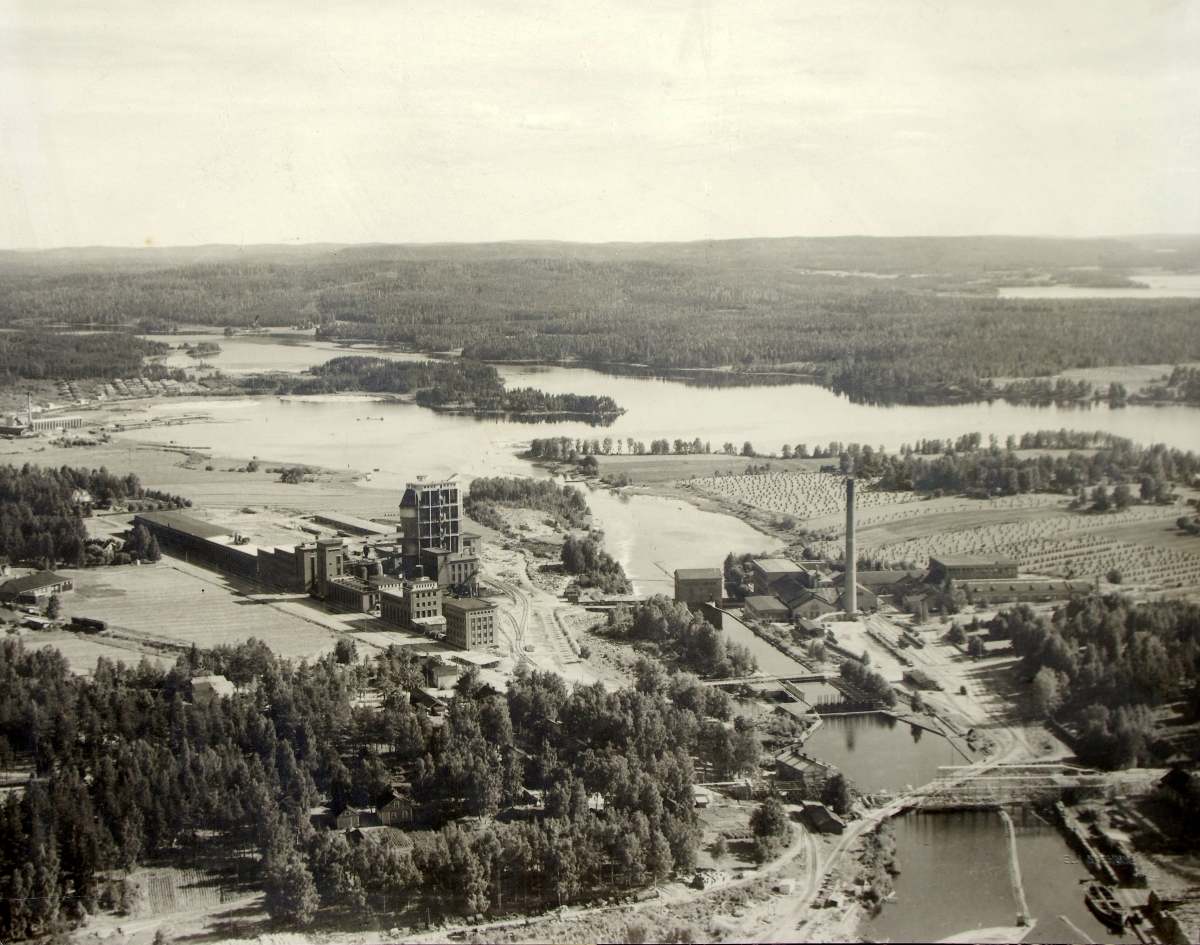Hiskinmäen maantiesilta on valmistunut 1950, joten kuva on aivan 1940-luvun lopusta. Ennen uuden sillan valmistumista hiskinmäkeläiset kulkivat koskiuoman yli viistoon kulkevaa Punaista siltaa pitkin. Vasemmassa yläreunassa näkyy yhtiön uudemman sahan rakennuksia Kuhnamon rannalla ja siitä alaspäin sulfiittiselluloosatehtaan ja kemiallisen rakenuksia. Piilolanniemen pelloilla on heinää pantu seipääille, joten kuva on todennäköisesti heinäkuulta 1949.