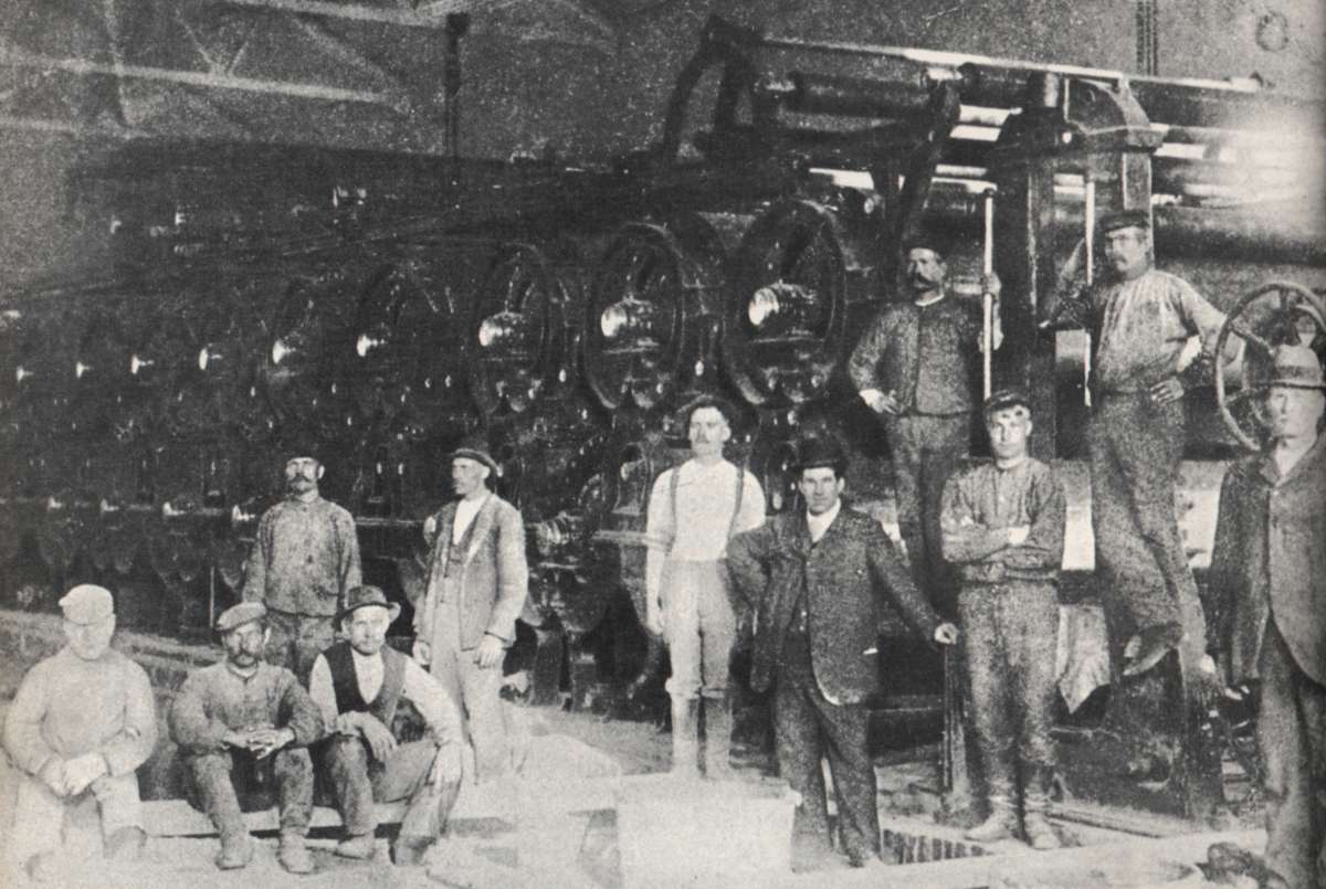 Kuva on tiettävästi otettu uuden paperikoneen asennustyön valmistumisen kunniaksi vuonna 1906. Kuvasta tunnistetaan ainakin äärimmäisellä vasemmalla Oskar Tanttinen ja äärimmäisenä oikealla Edvald Ahlsten. Paperikoneen edessä seisoo ensimmäisenä vasemmalta Fabian Rinne ja kolmantena Sylvester Koskinen. Hattupäinen kaiteeseen nojaava herrasmies on amerikkalainen asentaja, jonka nimi on ikävästi unohtunut.