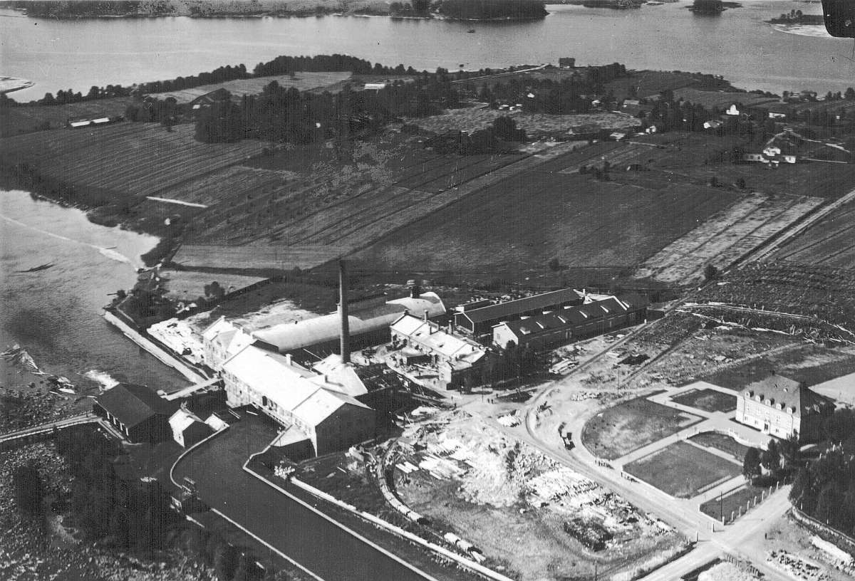 Paperi- ja kartonkitehtaan ympäristöä vuodelta 1925. Tuolloin yhtiön höyrykattilalaitosta uudistettiin ja samalla voimalaitokselle rakennettiin uusi 78-metriä korkea savupiippu. Kuvassa rakennustyöt ovat vielä kesken. Yhtiön vuonna 1923 valmistunut pääkonttori on jo valmiina. Kuvan yläosassa hieman keskeltä oikealla Kuhnamon rannassa näkyy Kalle Piilosen höyrysahan konttorirakennus. Nimismies Erkki Seppä osti rakennuksen myöhemmin asunnokseen, josta johtuen koko aluetta alettiin kutsua Nimismiehenrannaksi.