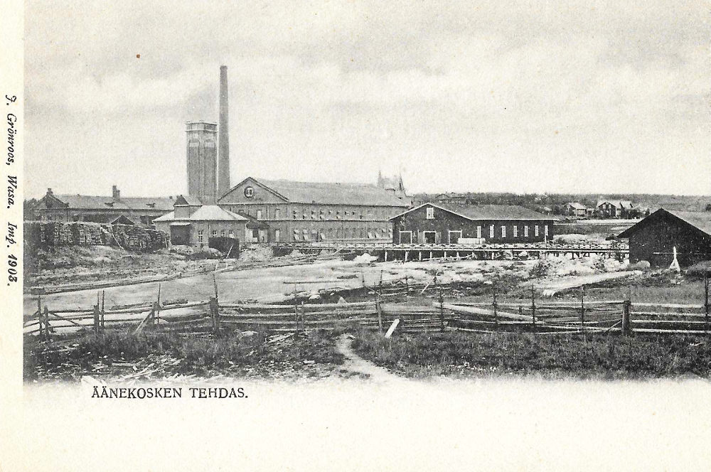Äänekoski Oy:n puumassa- ja kartonkitehdasta aivan 1900-luvun alussa kosken itärannalta kuvattuna. Kuvan keskellä on kaksikerroksinen kartonkikonehalli sekä sen takana näkyy höyryvoimalaitos savupiipuineen. Savupiipun vieressä on 30-metriä korkea massa-arkkien kuivaukseen käytetty kuivauskanaali. Kuvan vasemmassa reunassa näkyy höyryvoimalaitoksen puulaanin sivua. Kartonkitehtaan tuotanto kuljetettiin maailmalle koskiuoman yli kulkevaa kapearaiteista rautatietä pitkin. Tämä kuva on otettu ennen vuotta 1926, jolloin höyryvoimalaitokselle rakennettiin laajennus ja samalla muurattiin korkeampi 78-metrinen savupiippu kuvassa näkyvän vanhan savupiipun etupuolelle. Kartonkitehdashallin takana näkyy Klubirakennuksen tornimaista kattoa ja siitä oikealla kansanopiston rakennus aivan koskiuoman varrella. Kosken itärannalla oli vielä tähän aikaan Mannilan peltoja ja niittyjä risuaitoineen.